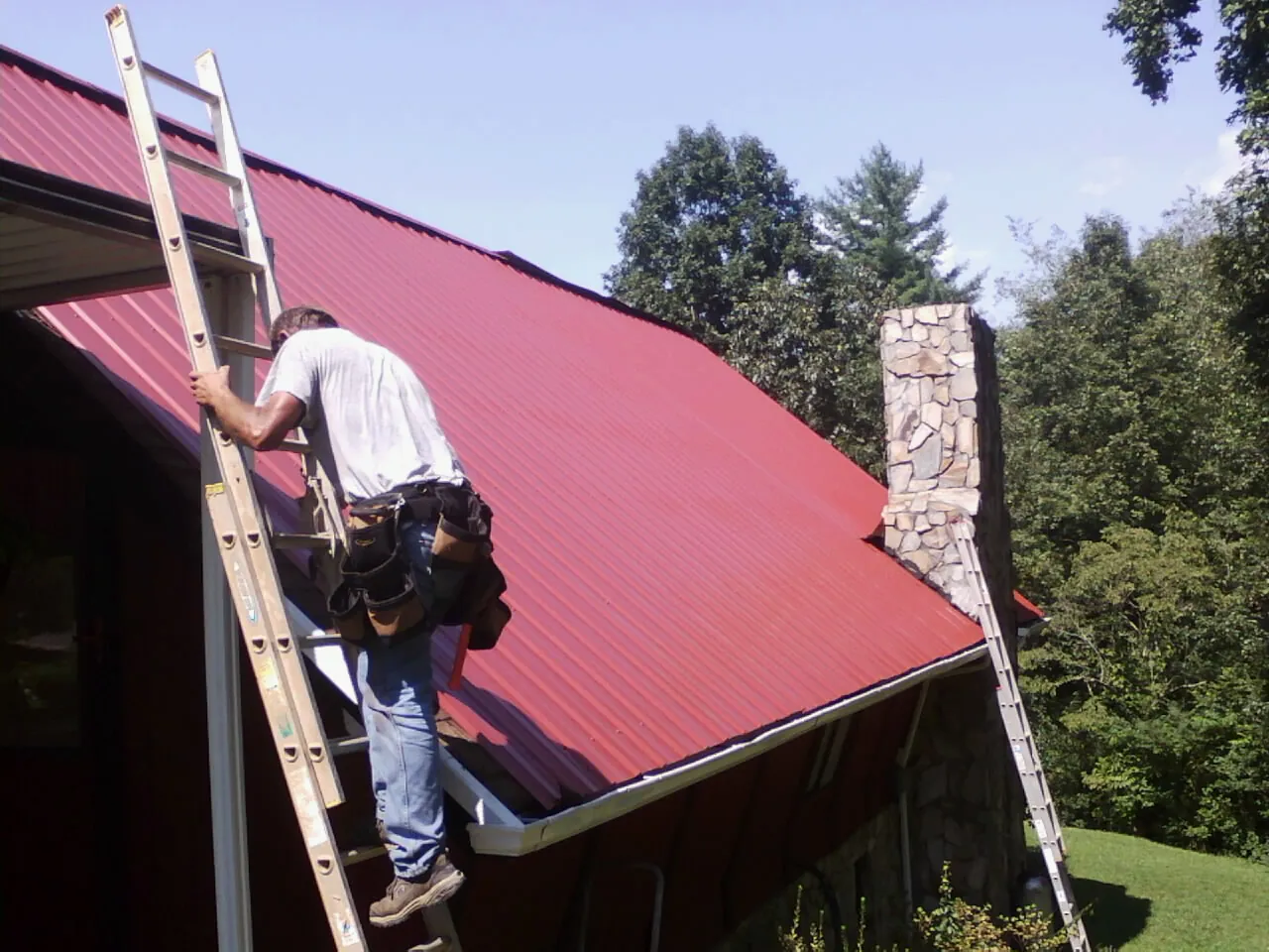 Roofer on a ladder beside a red metal roof and stone chimney — Soffit & Fascia Repair by Roofing Brothers in Delmar